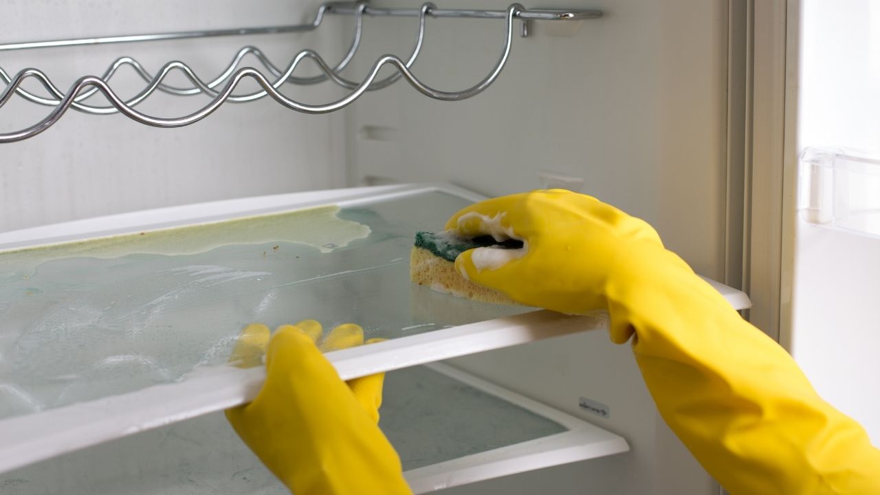 Tita Kleans deep cleaning a fridge using a sponge while wearing protective yellow gloves.
